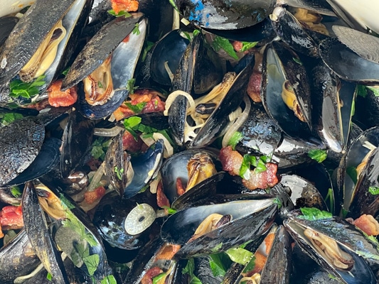 Close up shot of steamed mussels tossed with pieces of smoky bacon, sliced shallots, slices of garlic, and fresh green herbs.