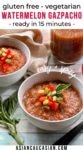 Two white bowls filled with spicy watermelon gazpacho on a wooden board, with ingredients on the side: a tomato, basil, and a spoon.