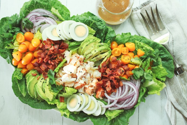 A platter of vibrant lobster cobb salad on top of romaine greens and a large fork and clear jar of miso dressing on the side.