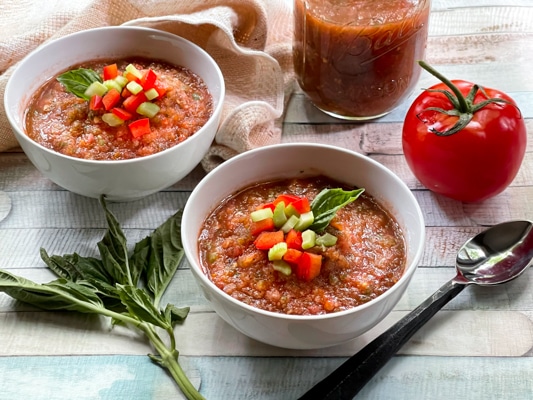 Two white bowls filled with spicy watermelon gazpacho on a wooden board, with ingredients on the side: a tomato, basil, and a spoon.