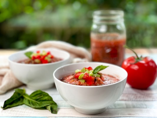 Two white bowls filled with spicy watermelon gazpacho on a wooden board, with ingredients on the side: a tomato, basil, and a clear jar of gazpacho.