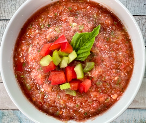 A white bowl filled with spicy watermelon gazpacho topped with fresh chopped veggies on a wooden board.