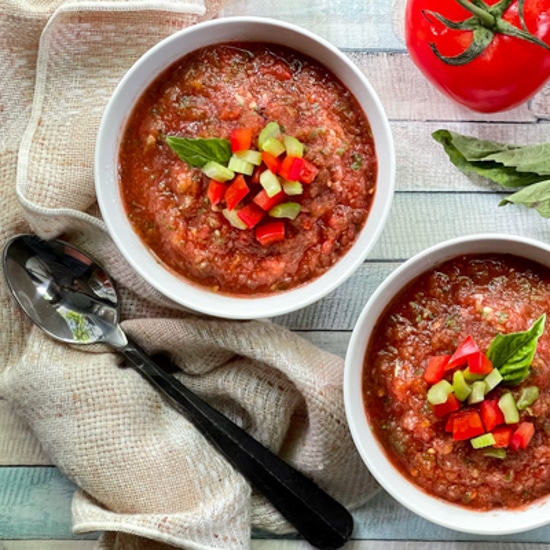 Two white bowls filled with spicy watermelon gazpacho on a wooden board, with ingredients on the side: a tomato, basil, basil, and a spoon.