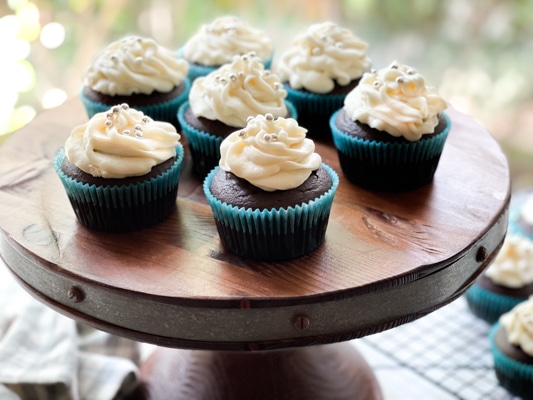 A close-up image of a chocolate miso cupcake with white frosting topped with silver sprinkles, on a wooden board.