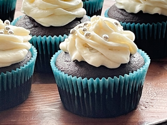 A close-up image of a chocolate miso cupcake with white frosting and silver sprinkles, on a wooden board.