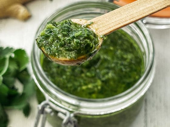 A clear glass bowl of cilantro, mint, basil sauce with a small wooden spoon inserted, and bunches of fresh cilantro and basil leaves, and ginger root, all on top of a white wooden board.