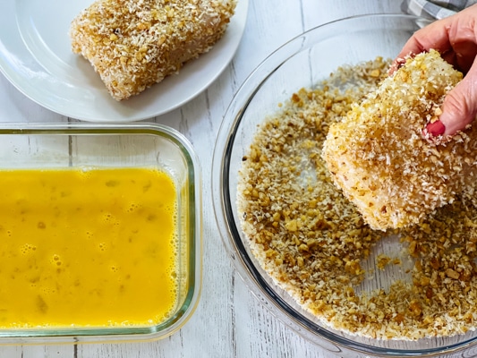 A woman's hand dredging a piece of halibut into panko bread crumbs, with a bowl of whisked eggs on the side.