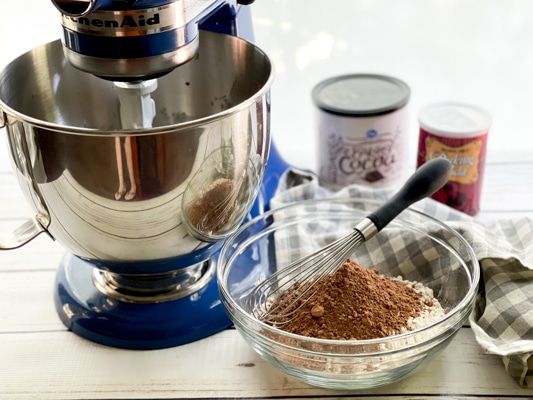 A clear mixing bowl filled with flour and cocoa powder and a whisk on top, and a blue Kitchen Aid mixer on the side, with boxed ingredients in the background.