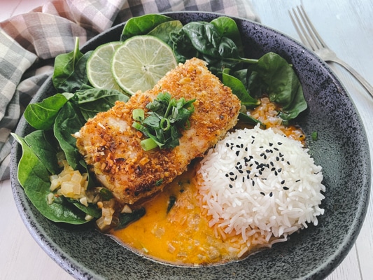 A golden crusted halibut fillet in a black bowl with a vibrant red curry sauce at the bottom of the bowl, and white steamed rice and spinach on the side.