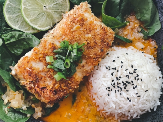 A beautiful piece of golden crusted halibut fillet in a black bowl with a vibrant red curry sauce at the bottom of the bowl, and white steamed rice and spinach on the side.