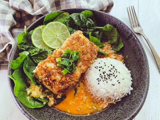 A beautiful piece of golden crusted halibut fillet in a black bowl with a vibrant red curry sauce at the bottom of the bowl, and white steamed rice and spinach on the side.