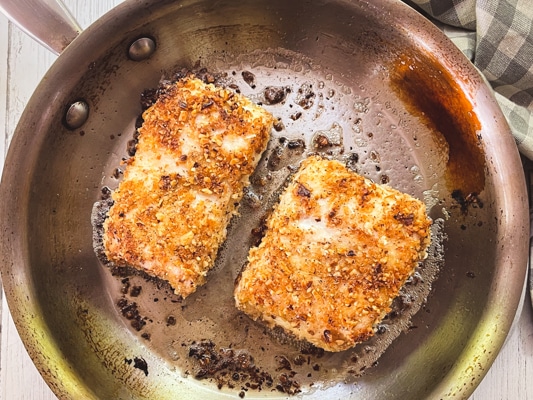 Two pieces of golden brown halibut fillets being seared in a stainless steel frying pan.