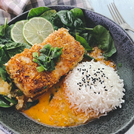 A beautiful piece of golden crusted halibut fillet in a black bowl with a vibrant red curry sauce at the bottom of the bowl, and white steamed rice and spinach on the side.