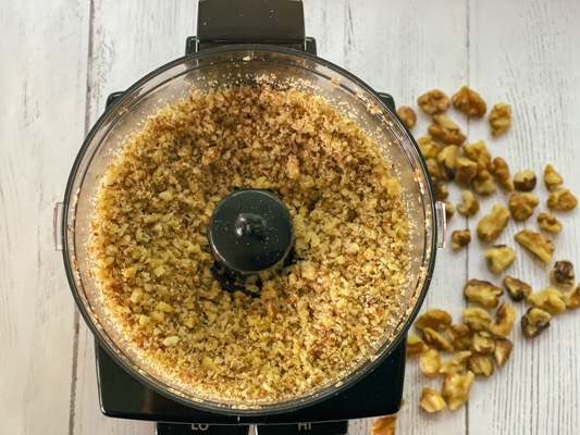 Chopped walnuts in a food processor with pieces of walnuts on a white board.