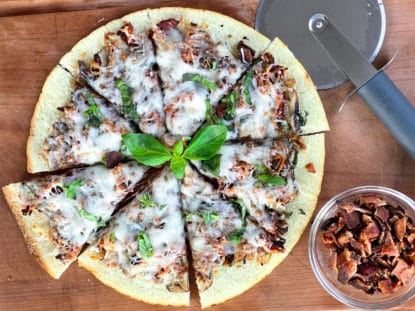 A sliced cauliflower pizza pie topped with bacon, shiitake mushrooms, basil, and melty cheese, with bacon bits on the side and a pizza cutter, on top of a wooden board.