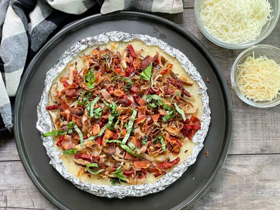 An uncooked cauliflower pizza pie topped with bacon, shiitake mushrooms, and basil with two cheeses on the side on top of a wooden board.