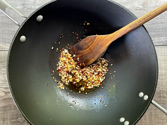 Garlic and red pepper flakes frying in a black wok with a wooden spatula inserted.