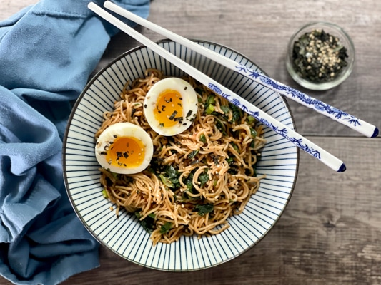 A striped bowl filled with ramen noodles topped with two runny eggs, with chopsticks laying on top of the bowl.