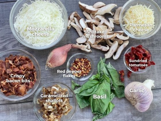 Ingredients on a board for making cauliflower pizza, including bacon bits, caramelized shallots, two cheeses, basil, shiitake mushrooms, sundried tomatoes, and basil.