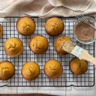 Freshly baked pumpkin muffins on a wire baking rack with a woman brushing the tops with butter.
