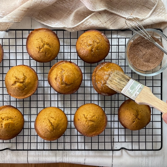 Freshly baked pumpkin muffins on a wire baking rack with a woman brushing the tops with butter.