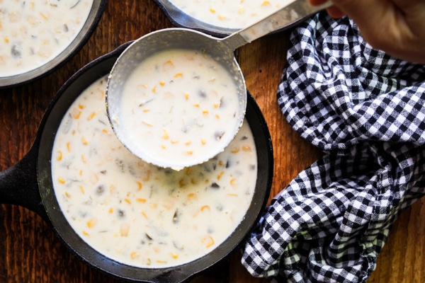 Batter for making skillet jalapeño corn bread in a large glass mixing bowl with a ladle inserted, and in two small cast iron skillets, on top of a wooden board with a black and white checkered napkin on the side.