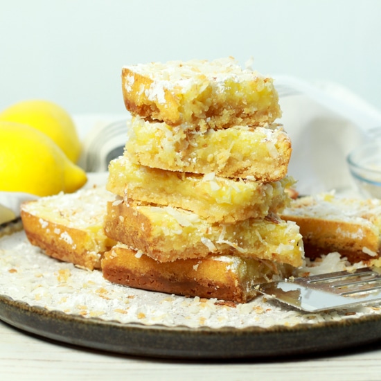 A stack of gluten-free lemongrass coconut squares on a white plate with fresh lemons in the background.
