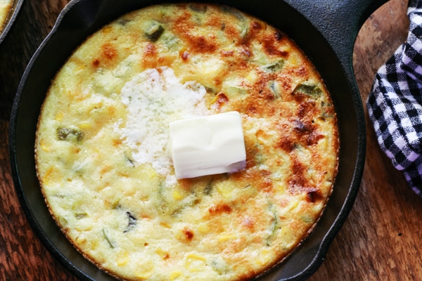 A mini skillet of baked jalapeño corn bread with a pat of butter on top and a black and white checkered napkin on the side, on top of a wooden board.