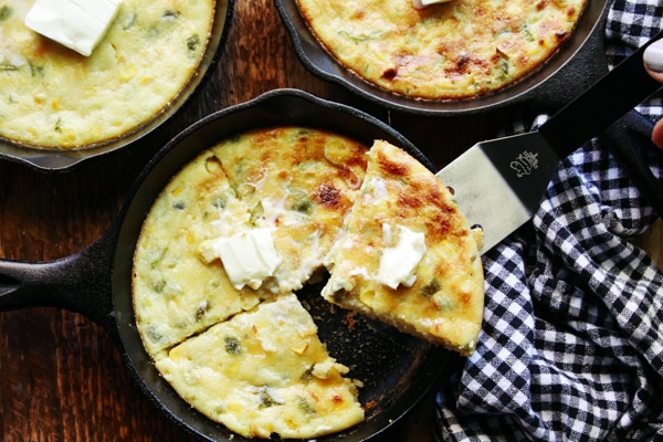 A piece of skillet jalapeño corn bread being lifted out of a mini skillet with a pat of butter on top and a black and white checkered napkin on the side and two other mini skillets behind.