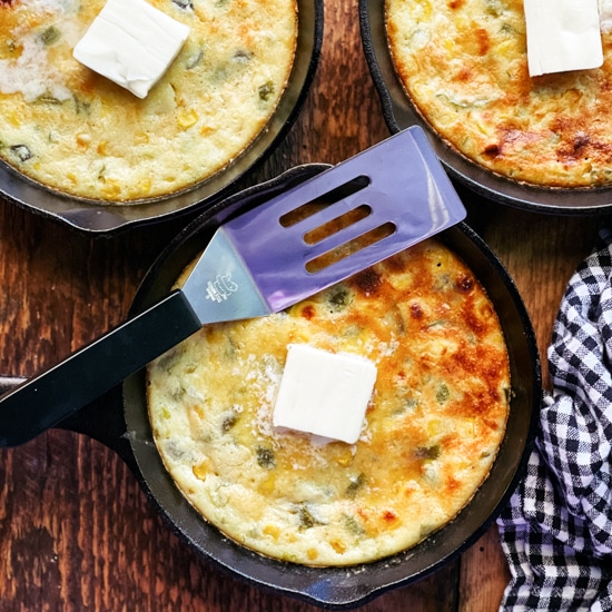 Three mini skillets of baked jalapeño corn bread with a pat of butter placed on top and a spatula and black and white checkered napkin on the side, on top of a wooden board.
