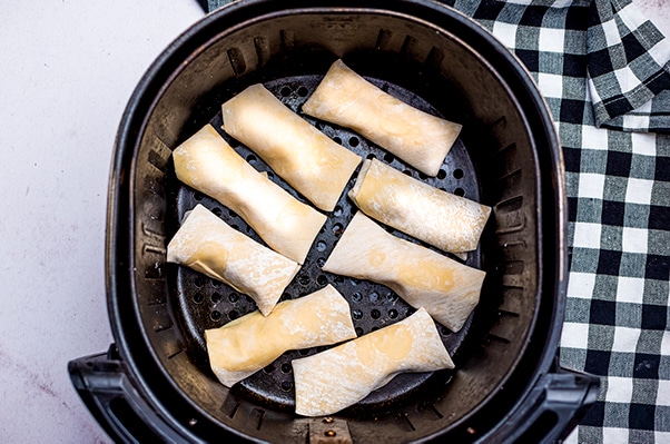 Uncooked vegan avocado egg rolls in an air fryer tray, ready for frying with a black and white checkered napkin on the side.