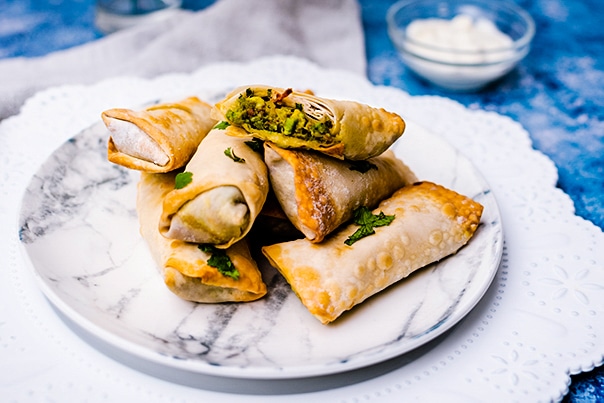 Vegan Avocado Egg Rolls stacked on a white marbled plate on top of a blue board with a small bowl of sour cream in the background.