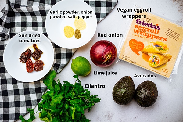 Ingredients for making vegan avocado eggrolls on a white board with a black and white checkered napkin on the side.