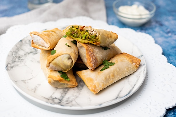 Vegan Avocado Eggrolls stacked on a white marbled plate on top of a blue board with a small bowl of sour cream in the background.