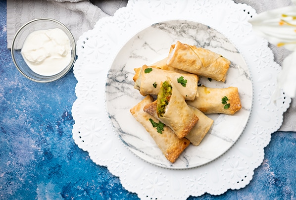 Vegan Avocado Eggrolls stacked on a white marbled plate on top of a blue board with sour cream on the side.