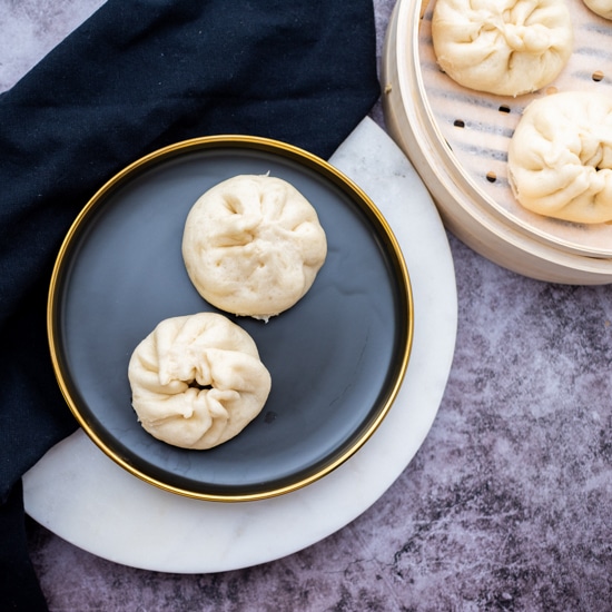 Two Chinese steamed pork buns on a round black plate with a bamboo steamer on the side filled with steamed buns, on top of a gray marbled surface.
