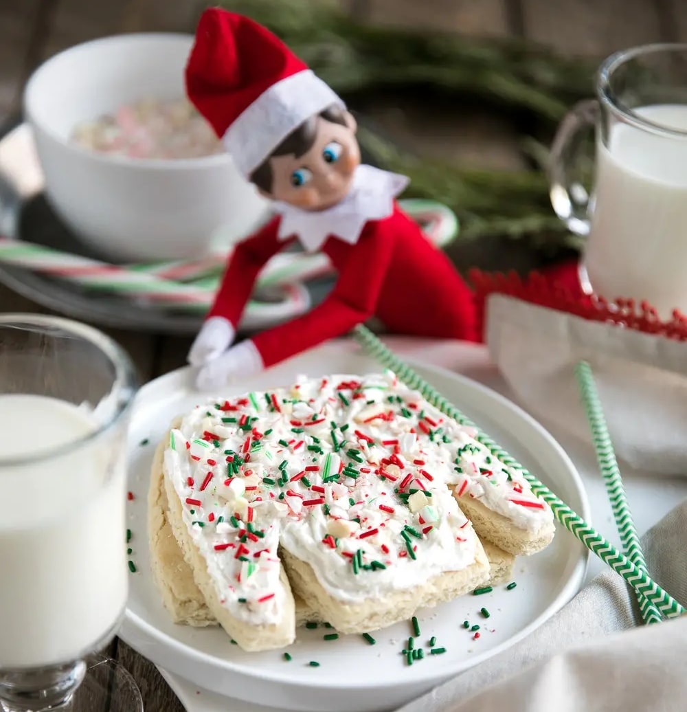 A tiny elf hovering over a plate of ugly sweater sugar cookies with a side of milk on the side.