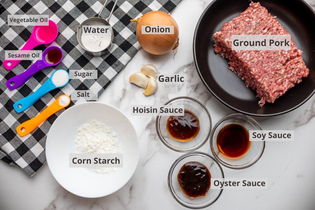 Various labeled ingredients in bowls for making the filling for Chinese steamed pork buns, on top of a marble surface with four colorful spoons on the side.