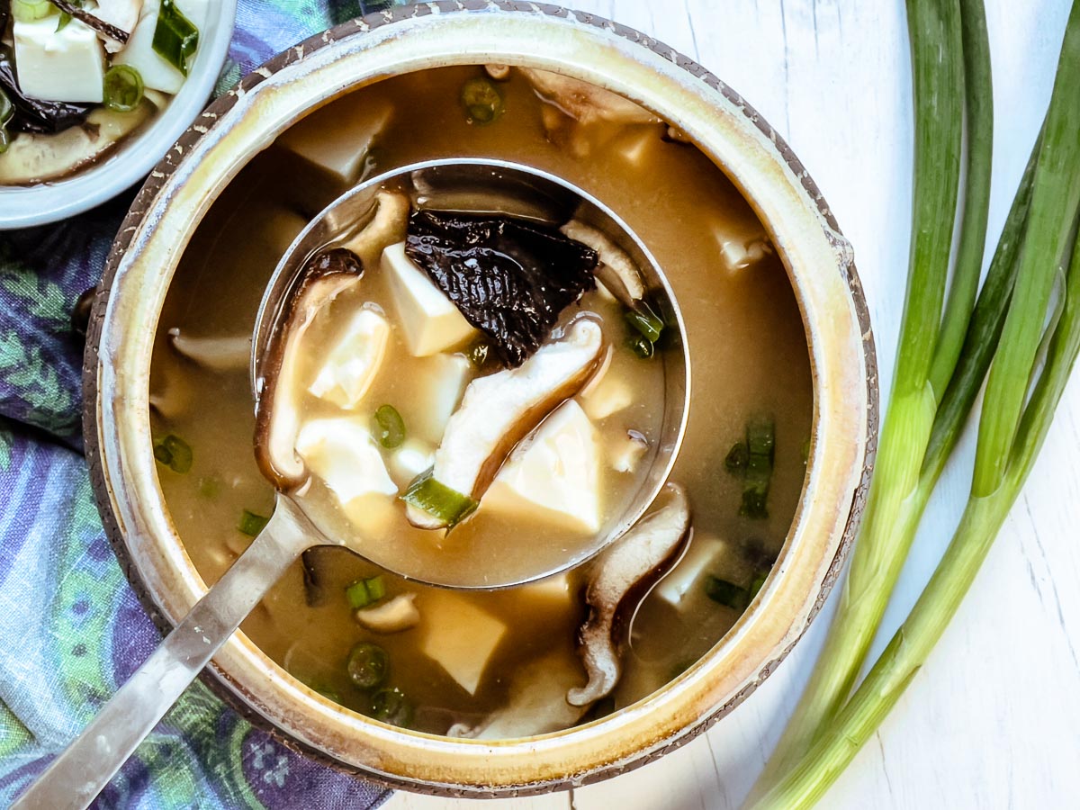 A soup bowl with a ladle spooning out miso soup with tofu and shiitake mushrooms with green onions on the side.