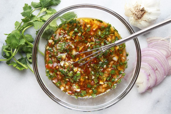 A glass bowl with Nuoc Cham dressing and a small whisk with sliced red onion, cilantro, and garlic on the side.