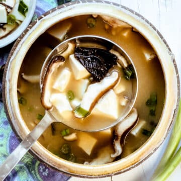 A soup bowl with a ladle spooning out miso soup with tofu and shiitake mushrooms with green onions on the side.