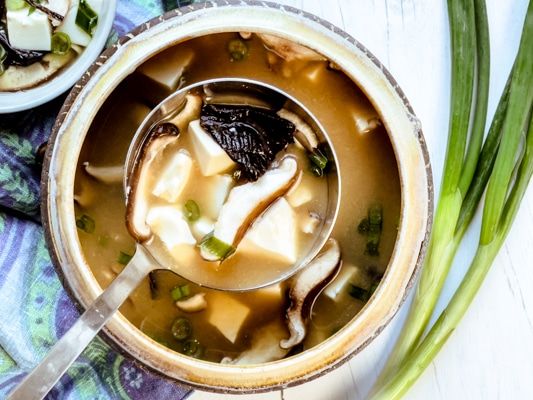 A soup bowl with a ladle spooning out miso soup with tofu and shiitake mushrooms with green onions on the side.