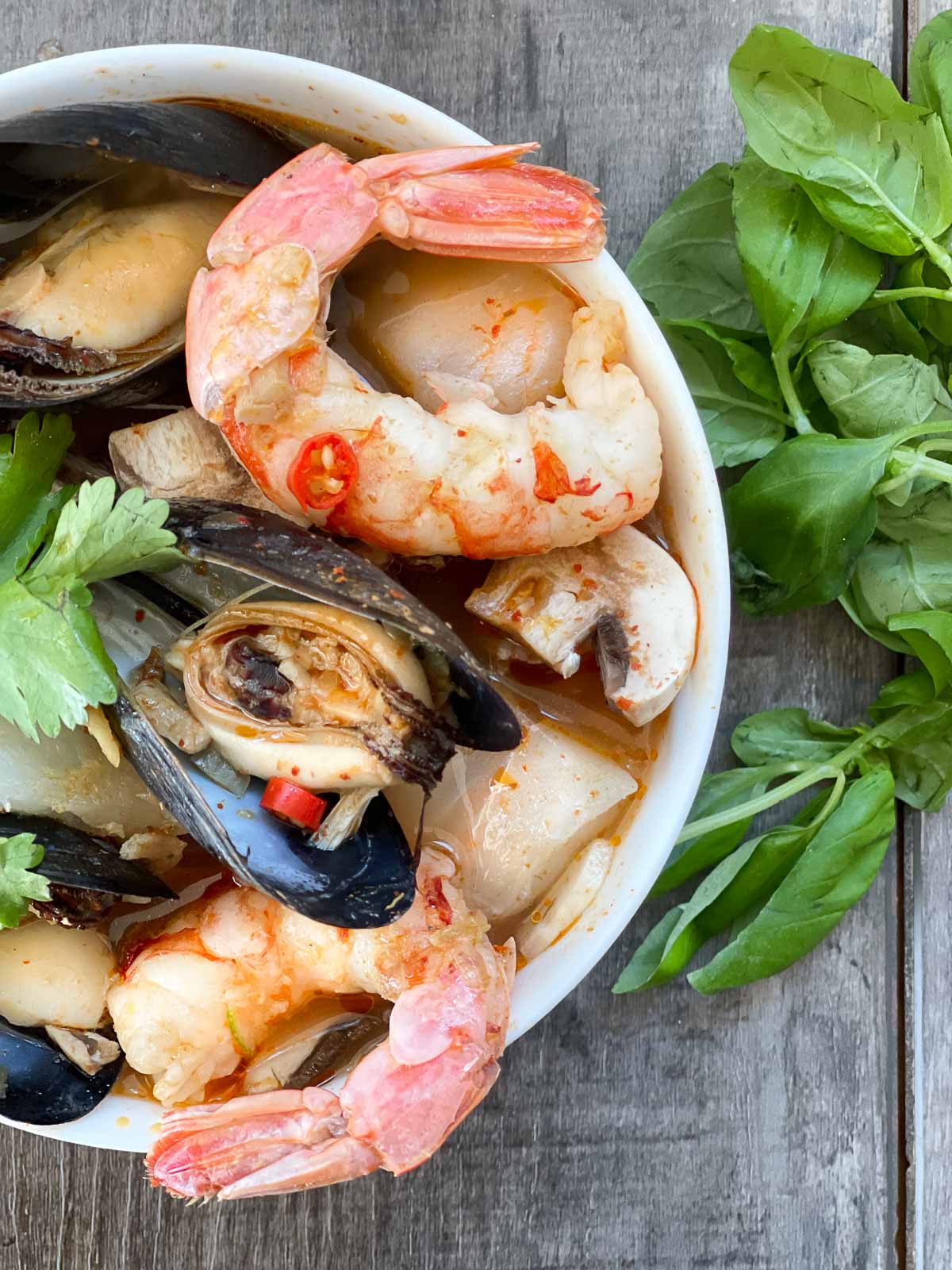 A white bowl of Thai Coconut Curry Seafood Soup with shrimp, mussels and fish with fresh basil on the side placed on top of a wooden board.