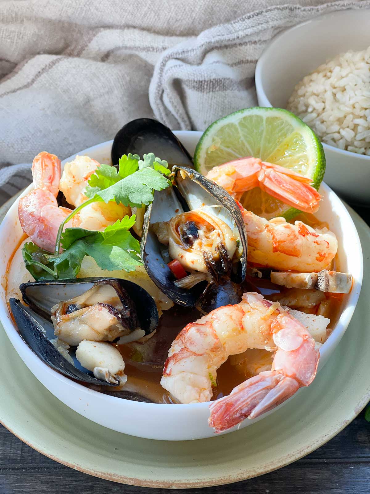 A white bowl of Thai Coconut Curry Seafood Soup with shrimp, mussels and lime on top of a yellow plate and a small bowl of rice on the side placed on top of a wooden board.