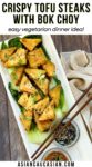 Golden-brown, crispy tofu steaks piled on top of cooked baby bok choy and sprinkled with seasonings plated on top of a white serving platter with small bowl of sauce and furikake seasoning on the side, and a pair of chopsticks on top and a napkin on the side.