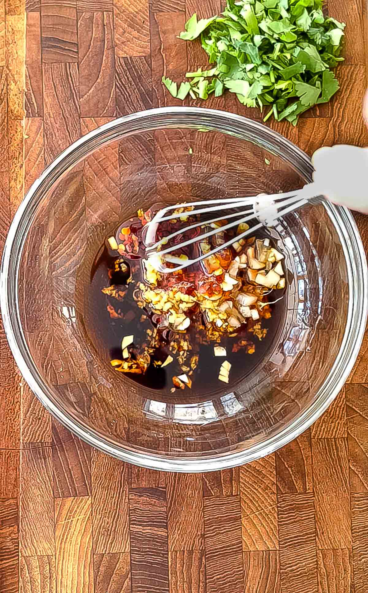 A glass bowl of chili oil being stirred with a whisk on top of a wooden board with fresh chopped cilantro on the side.