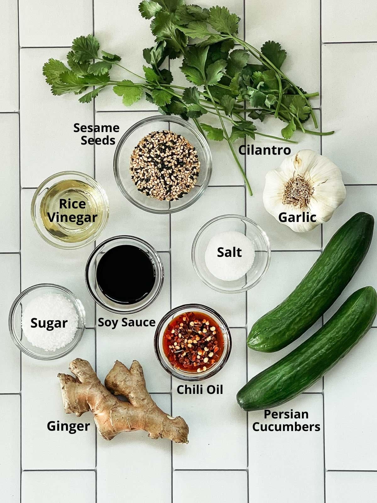 Labeled ingredients on a white tile surface for making smashed cucumber salad.