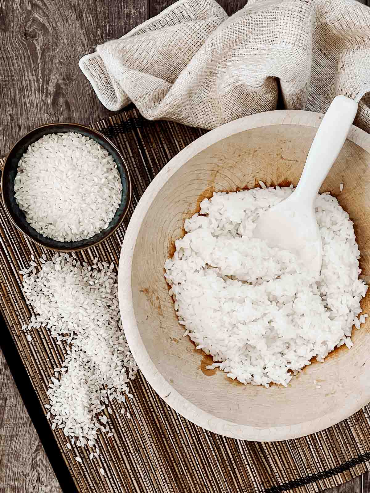 A wooden bowl of steamed sushi rice with a white paddle inside and a small bowl of uncooked rice on the side with a napkin on top of a bamboo mat.