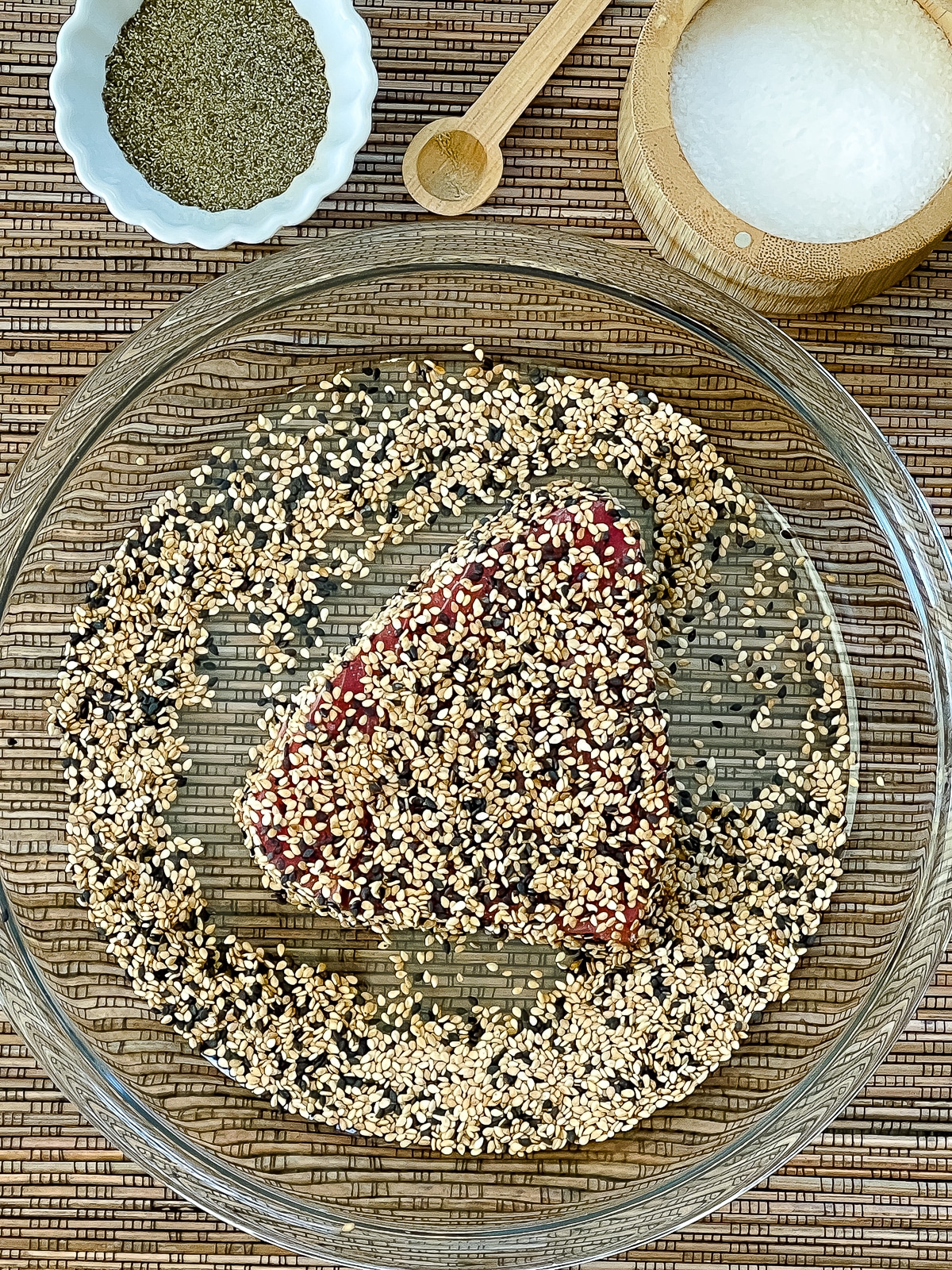 A tuna steak coated in sesame seeds being dredged in a glass bowl with two bowls of salt and pepper placed behind on top of a bamboo mat.
