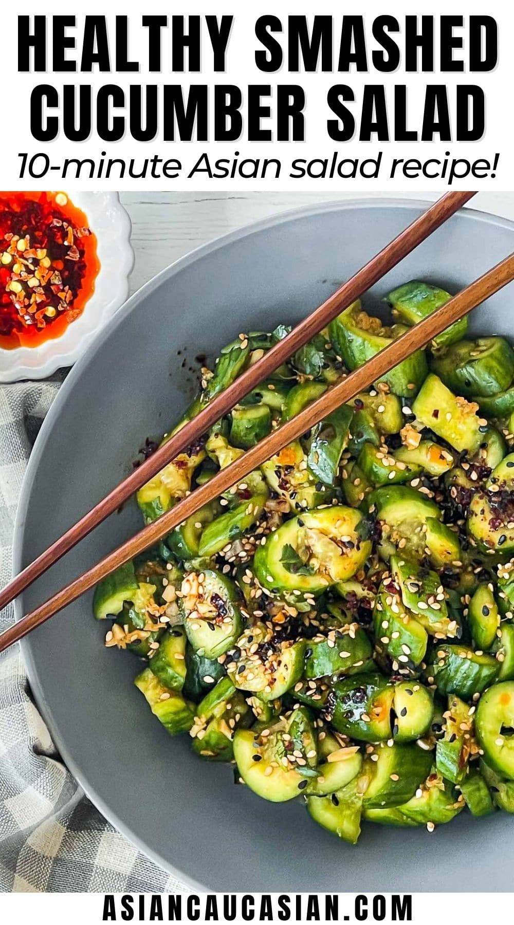 Smashed cucumber salad in a gray bowl with wooden chopsticks on top and a small white bowl of chili oil and a checkered napkin on the side.